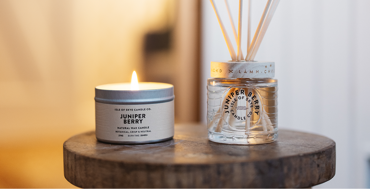 A lit candle and reed diffuser by the Isle of Skye Candle Company are sat onto of a dark wood stool in a warm lit home.