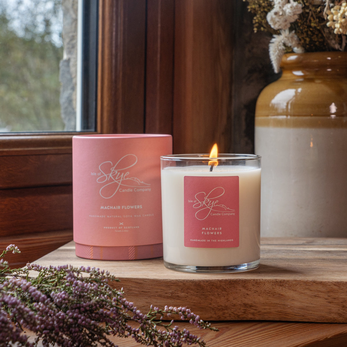 Isle of Skye Candle Company's candle, Machair Flowers, sitting onto of a plank of wood. A vase with flowers features in the background and is sat next to a window.